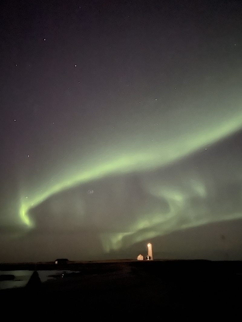 Green ribbons of northern lights arc above a small illuminated lighthouse on the Icelandic coast.