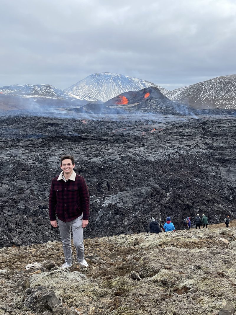 Maximilian Gebauer standing on a young lava field with an active volcanic fissure erupting behind him in Iceland.